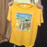 El color base de la camiseta es amarillo. La imagen muestra a dos boricuas que transmiten la tradición de la identidad puertorriqueña, especialmente a través de la vida yaucana. Combina nuestra vida local con las tradiciones puertorriqueñas nacionales.